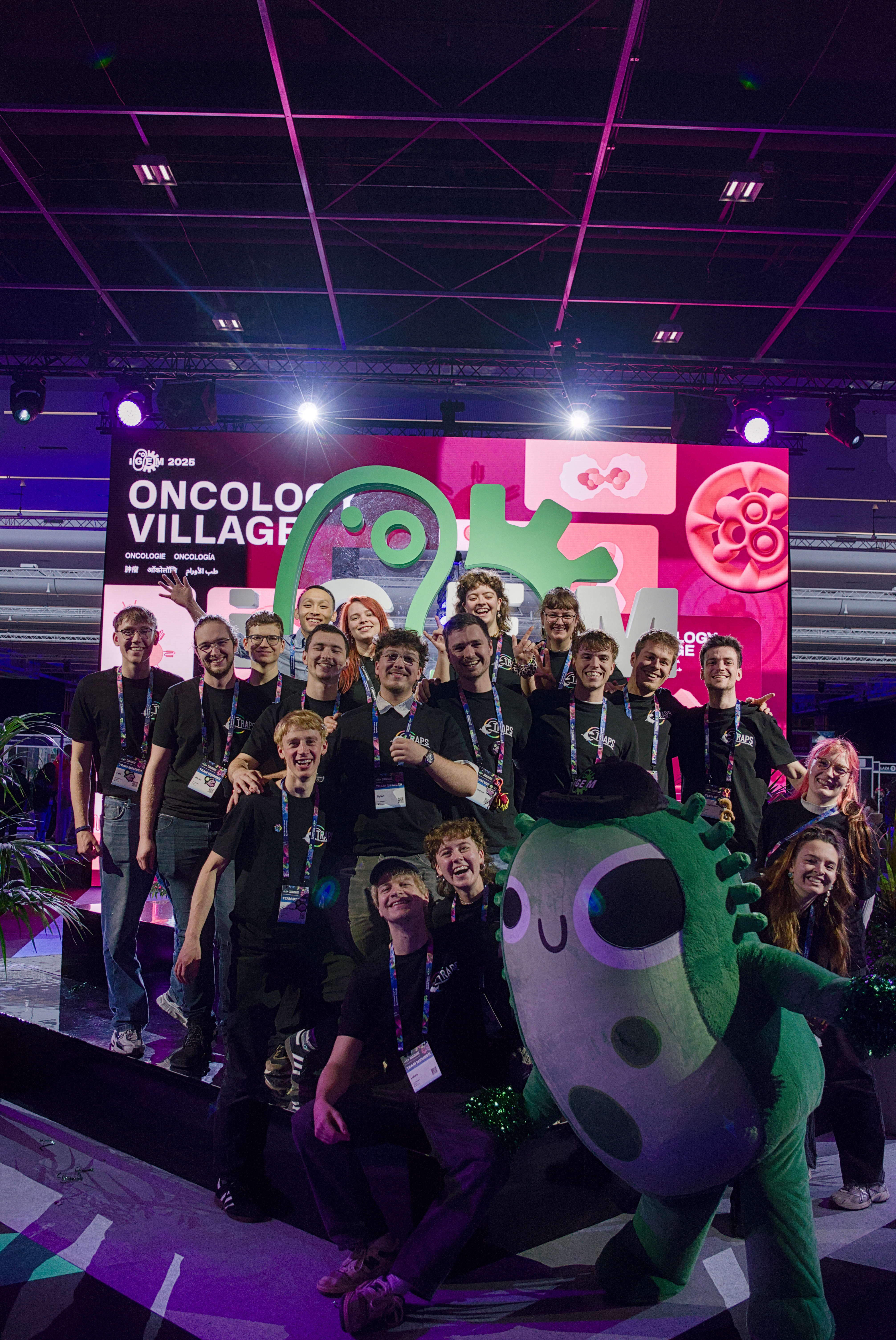 TRAPS posing in front of the iGEM sign, alongside iGEM' mascot: Gemy, a green microbe.