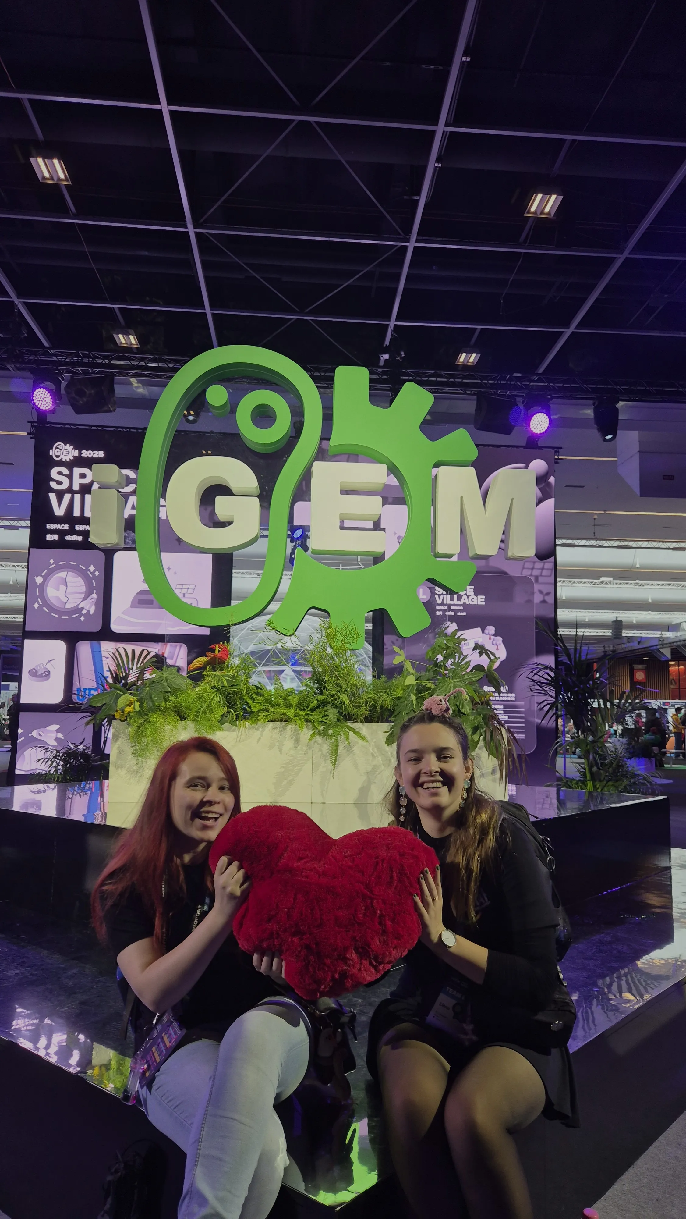 Two girls holding up a red plush heart in front of the iGEM sign