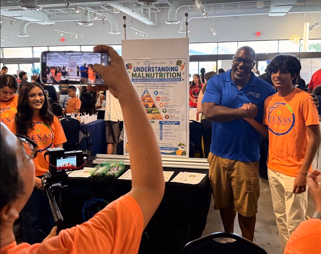 Team members in Texas iGEM shirts talking with a visitor at a table carrying the Understanding Malnutrition chart and the $3 Meal Plan recipe cards.