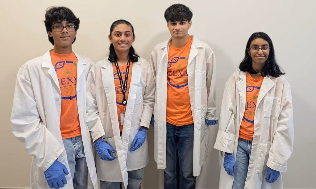 Four students in white lab coats, orange Texas iGEM shirts and blue nitrile gloves, standing side by side against a plain wall.
